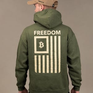 May include: An olive green hooded sweatshirt featuring a cream-colored graphic on the back. The graphic displays the word FREEDOM above a stylized flag design incorporating a Bitcoin symbol within a square. A tan baseball cap is worn on the head.