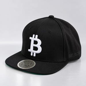 May include: A black baseball cap featuring a white embroidered Bitcoin symbol on the front panel. The word MERCH is embroidered on the side, and a circular silver sticker with a maze-like logo is affixed to the flat brim, which has a green underside.