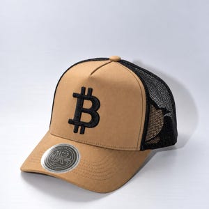 May include: A tan baseball cap with a black mesh back and a black embroidered Bitcoin symbol on the front panel. The cap features a circular silver sticker on the brim with the text MERCH and a stylized logo design, including a button on top.