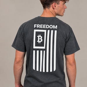 May include: A dark charcoal gray short-sleeve t-shirt featuring a white graphic on the back. The design displays the word FREEDOM above a stylized flag graphic that incorporates a Bitcoin symbol within the rectangular canton area.
