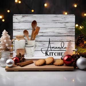 Peut inclure: Une planche à découper en bois blanc avec une finition vieillie. La planche présente un design rustique avec un sapin de Noël et le texte "Amanda Kitchen". La planche est décorée de cuillères en bois, d'un bocal et d'une tasse. La planche est posée sur un plateau en bois avec des bâtonnets de cannelle et des biscuits.