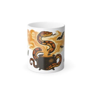 May include: White ceramic mug featuring a Halloween-themed design. The design includes a brown and orange snake rising from a black cauldron, with bats and ghosts in the background. The mug is a standard size for hot beverages.