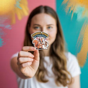 Op de afbeelding: Een sticker met een cartoon eekhoorn die noten vasthoudt, met de tekst "SQUIRREL BRAIN ACTIVATED!". De eekhoorn is bruin en oranje, met een pluizige staart. De sticker heeft een witte rand.