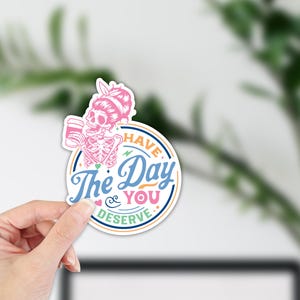 May include: A round sticker featuring a pink skeleton with a coffee cup and a pink bow. The sticker has the text "Have The Day You Deserve" in blue and white, with a pink and orange border.
