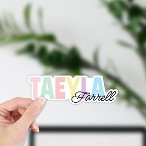 Puede incluir: Una pegatina con el nombre "TAEYLA Farrell" en colores pastel. La palabra "TAEYLA" está en letras de bloque, cada letra de un color diferente: rosa, verde, melocotón, amarillo, morado y azul. El nombre "Farrell" está en cursiva.