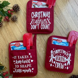 May include: Three red oven mitts with white text and designs. The first mitt says "Christmas calories don't count". The second mitt says "It takes a big heart to shape little minds". The third mitt says "Christmas baking crew".
