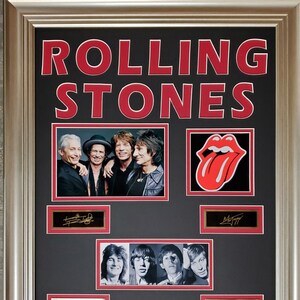 May include: Framed Rolling Stones memorabilia with the band's name in red. It features a group photo, the iconic tongue logo, and signatures. The frame has a silver tone.