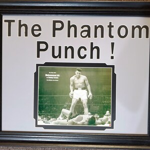 Può includere: Stampa incorniciata con cornice nera, raffigurante un'immagine in bianco e nero di Muhammad Ali su Sonny Liston. Il testo "The Phantom Punch!" è sopra l'immagine, con un passe-partout bianco.