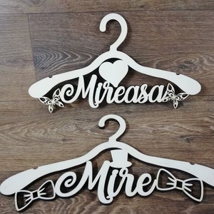 Può includere: Due grucce di legno bianche con ritagli decorativi. Una gruccia ha la scritta "Mireasa" con cuori e farfalle. L'altra ha "Mire" con cilindro e papillon. Su una superficie di legno.