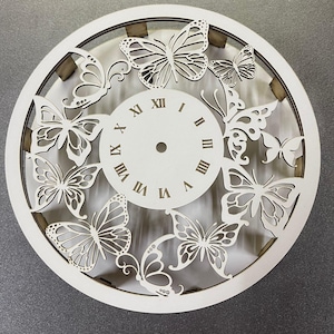 Puede incluir: Cara de reloj de madera blanca con números romanos y un diseño recortado de mariposas. La cara del reloj es redonda y tiene un diámetro de aproximadamente 30 cm.