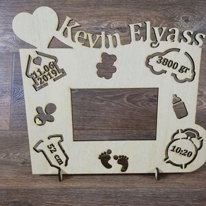Pode incluir: Moldura de fotos de madeira com um cora&ccedil;&atilde;o, uma casa, um urso de peluche, uma nuvem, uma mamadeira, um macac&atilde;o, pegadas e um despertador. A moldura &eacute; personalizada com o nome "Kevin Elyass", a data "11.06.2019", o peso "3800 g", o comprimento "52 cm" e a hora "10:20".