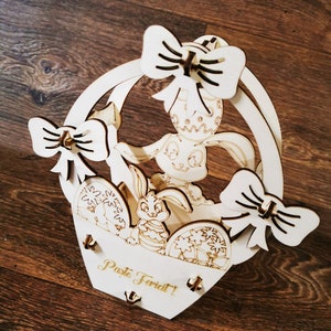 Puede incluir: Una cesta de Pascua de madera con un conejito y huevos decorados. La cesta tiene un asa y adornos de lazo. El texto "Paste Fericit!" es visible en la parte delantera. La cesta es de madera clara.