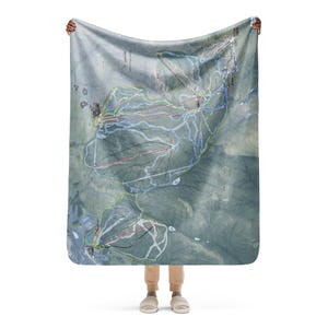 Può includere: Una grande coperta rettangolare con una mappa dettagliata di una stazione sciistica, con sentieri e altitudini segnati in vari colori. La coperta ha una palette di colori tenui di verdi, blu e marroni, con una leggera estetica vintage.