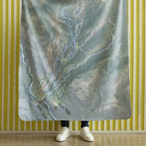 Puede incluir: Una manta rectangular grande con un mapa detallado de una estación de esquí, con senderos y remontes de varios colores. La manta se sostiene, mostrando el intrincado diseño del mapa. El fondo es una pared amarilla con rayas verticales.