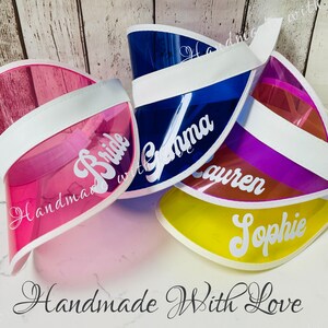 May include: Four clear plastic visors with white bands and personalised names in white script. The names are "Bride", "Gemma", "Lauren", and "Sophie". The visors are stacked on top of each other and the text "Handmade With Love" is written below the visors.
