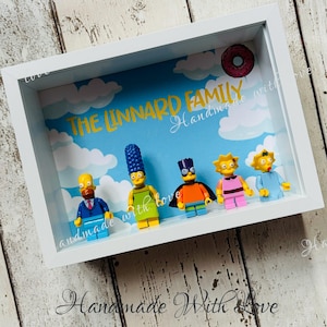May include: A white shadow box with a blue background and white clouds. The text "The Linnard Family Handmade with Love" is written in yellow. There are six yellow Lego minifigures inside the box, including Homer, Marge, Bart, Lisa, Maggie, and a donut.