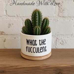 May include: A green cactus planted in a white ceramic pot with a light wood base. The pot features the black text "WHAT THE FUCCULENT" printed on the front. Above the plant, the text "Handmade With Love" is written in a cursive black font.