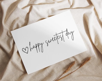 Op de afbeelding: Een witte kaart met de tekst "happy sweetest day" in elegant zwart schrift, vergezeld van een klein hartje. De kaart ligt op een beige stoffen ondergrond, met twee tarwearen in de rechter benedenhoek.