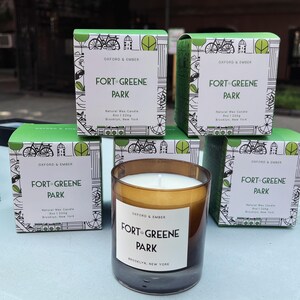 Puede incluir: Una vela de vidrio color ámbar con una etiqueta blanca que dice "FORT GREENE PARK" y "BROOKLYN, NEW YORK". Está rodeada de cajas verdes con el mismo texto y una ilustración de bicicleta. Las cajas también dicen "OXFORD & EMBER".