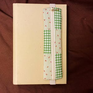 Puede incluir: Un libro color crema con un marcapáginas decorativo. El marcapáginas tiene una cremallera y está hecho de tela con cuadros verdes y patrones de cerezas rojas. El marcapáginas está sujeto a la parte superior del libro.