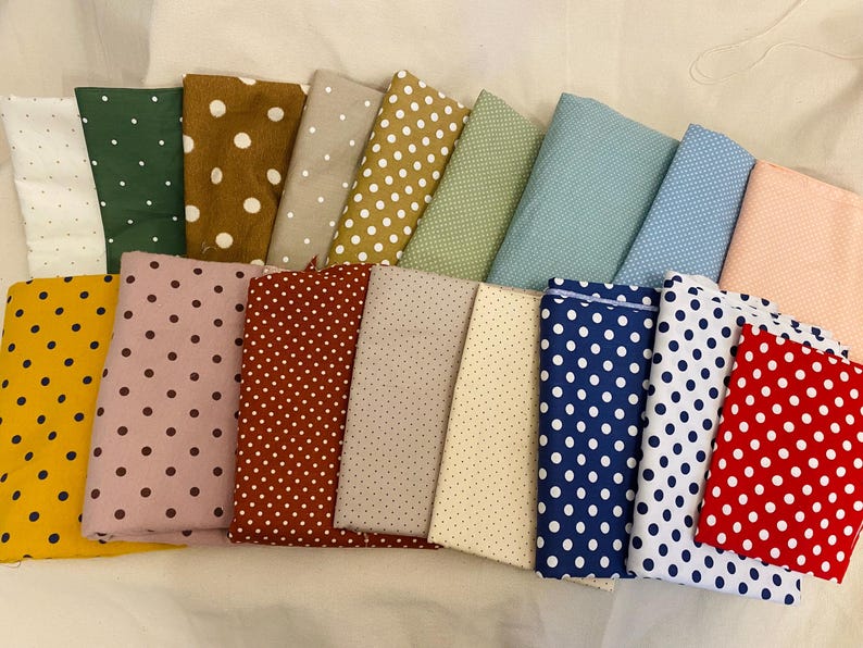 K&ouml;nnte beinhalten: Eine Sammlung gefalteter Stoffst&uuml;cke in verschiedenen Farben, jeweils mit einem Tupfenmuster versehen. Zu den Farben geh&ouml;ren Wei&szlig;, Gr&uuml;n, Braun, Beige, Gelb, Rosa, Rot, Blau und Pfirsich. Die Stoffe sind in einer ansprechenden Anordnung angeordnet.