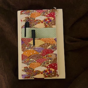 Könnte beinhalten: Ein Notizbuch mit einem farbenfrohen Stoffbezug mit einem japanischen Fächerdesign in Rot-, Orange- und Brauntönen. Zwei schwarze Stifte befinden sich in den Taschen. Das Notizbuch befindet sich auf einem cremefarbenen Hintergrund.