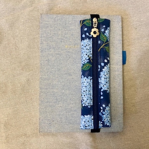 Peut inclure: Un carnet gris avec le mot "BULLET" en lettres dorées, associé à une trousse à crayons à imprimé floral bleu. La trousse a une fermeture éclair dorée et une tirette en forme de fleur. Le carnet a un signet ruban bleu.