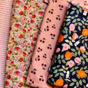 K&ouml;nnte beinhalten: Eine Auswahl an Stoffen mit verschiedenen Mustern. Ein Stoff hat rosa Streifen mit kleinen Punkten, ein anderer ein florales Erdbeerdesign, ein dritter ein Kirschmuster und der letzte ein Blumendesign mit orangefarbenen und rosa Bl&uuml;ten.