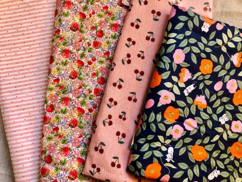K&ouml;nnte beinhalten: Eine Auswahl an Stoffen mit verschiedenen Mustern. Ein Stoff hat rosa Streifen mit kleinen Punkten, ein anderer ein florales Erdbeerdesign, ein dritter ein Kirschmuster und der letzte ein Blumendesign mit orangefarbenen und rosa Bl&uuml;ten.