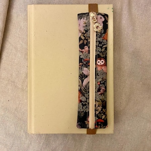 May include: A cream-colored book with a decorative bookmark. The bookmark features a black background with a colorful floral and animal print, including owls and mushrooms. A gold zipper and brown leather tab complete the design.