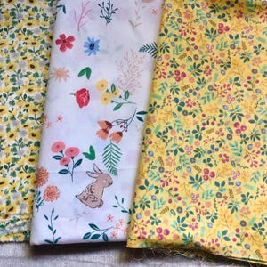 K&ouml;nnte beinhalten: Drei Stoffmuster mit floralen Mustern. Eines ist gelb mit kleinen wei&szlig;en und gelben Blumen. Ein weiteres ist wei&szlig; mit bunten Blumen und einer Hasenillustration. Das dritte ist gelb mit einer Mischung aus Blumen und Bl&auml;ttern.