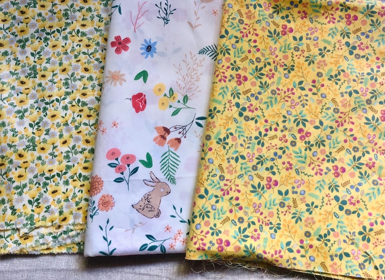 K&ouml;nnte beinhalten: Drei Stoffmuster mit floralen Mustern. Eines ist gelb mit kleinen wei&szlig;en und gelben Blumen. Ein weiteres ist wei&szlig; mit bunten Blumen und einer Hasenillustration. Das dritte ist gelb mit einer Mischung aus Blumen und Bl&auml;ttern.