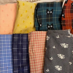 K&ouml;nnte beinhalten: Eine Sammlung gefalteter Stoffst&uuml;cke in verschiedenen Mustern und Farben. Designs umfassen rote Herzen, rosa Punkte, gelbe Tupfen, gr&uuml;nes Karomuster mit schwarzen Katzen und ein schwarz-wei&szlig;es Blumenmuster. Die Stoffe sind auf einer hellen Oberfl&auml;che angeordnet.