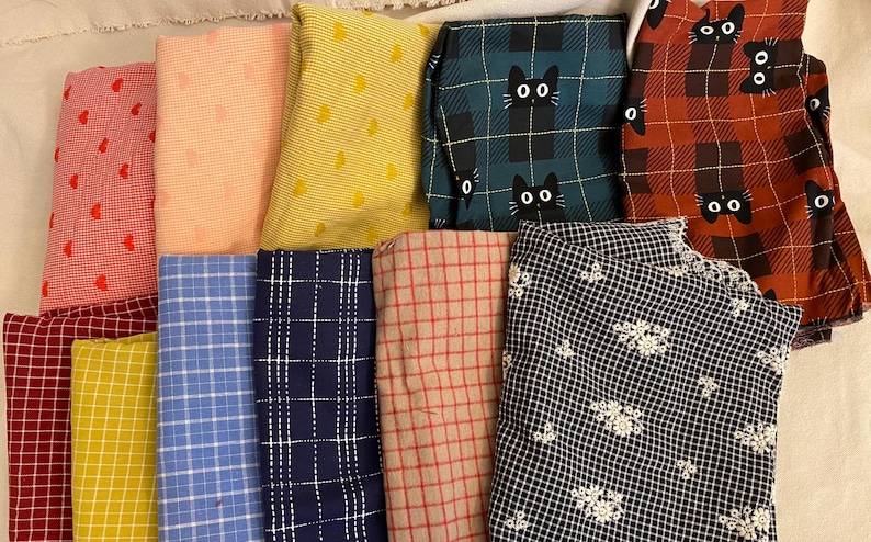 K&ouml;nnte beinhalten: Eine Sammlung gefalteter Stoffst&uuml;cke in verschiedenen Mustern und Farben. Designs umfassen rote Herzen, rosa Punkte, gelbe Tupfen, gr&uuml;nes Karomuster mit schwarzen Katzen und ein schwarz-wei&szlig;es Blumenmuster. Die Stoffe sind auf einer hellen Oberfl&auml;che angeordnet.
