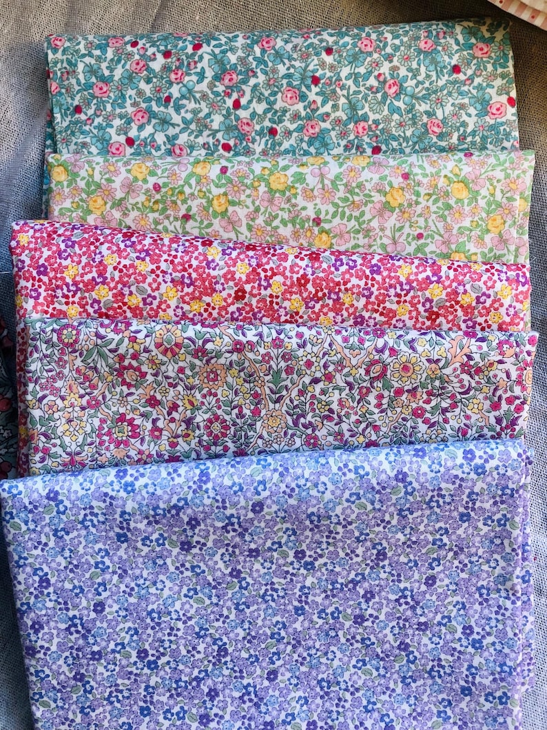 K&ouml;nnte beinhalten: Ein Stapel bunter Stoffe mit Blumenmustern. Die Stoffe zeigen verschiedene Muster kleiner Blumen in Rosa-, Rot-, Gelb-, Gr&uuml;n- und Lilat&ouml;nen. Die Stoffe sind ordentlich gefaltet und &uuml;bereinander gestapelt, was die Vielfalt der Designs zeigt.