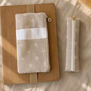 May include: A rectangular beige fabric pouch with a white daisy pattern and a horizontal white stripe, featuring a gold flower-shaped zip pull. Beside it is a slim, matching beige fabric pencil case with a white zip and gold hardware.