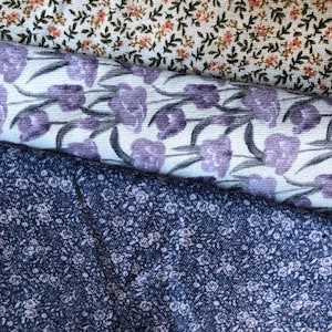 K&ouml;nnte beinhalten: Nahaufnahme von drei verschiedenen floralen Stoffmustern. Der obere Stoff ist cremefarben mit kleinen orangefarbenen und gr&uuml;nen Blumen. Der mittlere Stoff ist wei&szlig; mit lila tulpenartigen Blumen. Der untere Stoff ist marineblau mit winzigen wei&szlig;en Blumen.