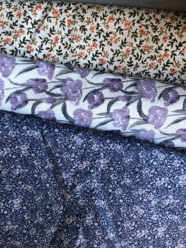 K&ouml;nnte beinhalten: Nahaufnahme von drei verschiedenen floralen Stoffmustern. Der obere Stoff ist cremefarben mit kleinen orangefarbenen und gr&uuml;nen Blumen. Der mittlere Stoff ist wei&szlig; mit lila tulpenartigen Blumen. Der untere Stoff ist marineblau mit winzigen wei&szlig;en Blumen.