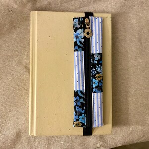 K&ouml;nnte beinhalten: Ein beiges Buch mit einem dekorativen Lesezeichen. Das Lesezeichen hat einen schwarzen Rei&szlig;verschluss und besteht aus Stoff mit blauen Blumen- und Streifenmustern. Ein goldfarbener Blumenanh&auml;nger ist am Rei&szlig;verschluss befestigt.
