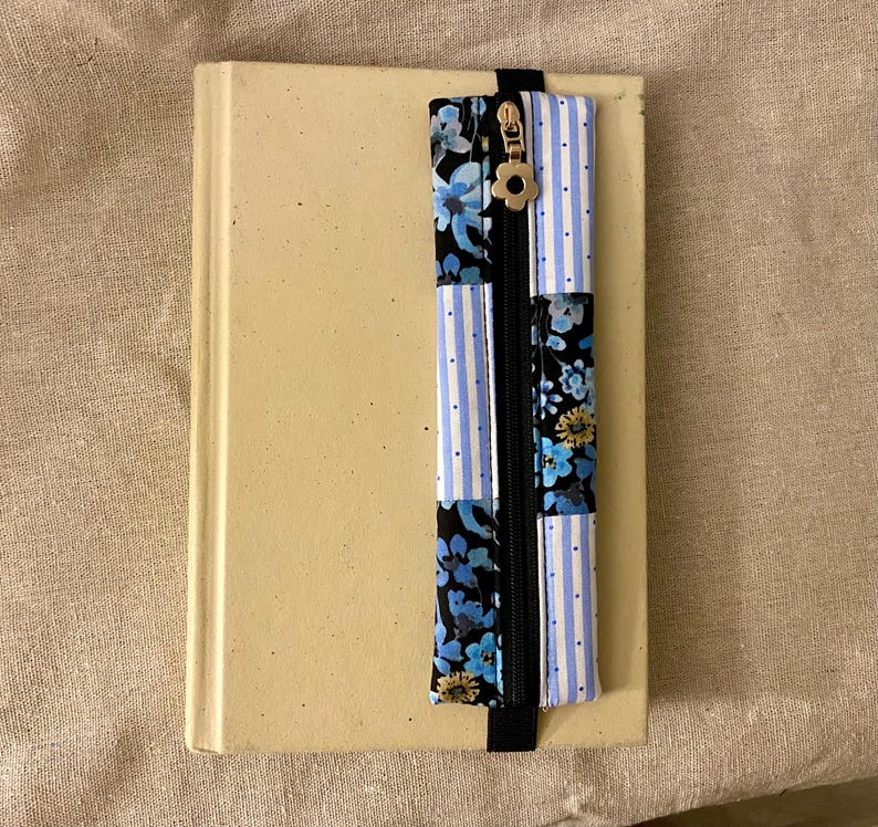 K&ouml;nnte beinhalten: Ein beiges Buch mit einem dekorativen Lesezeichen. Das Lesezeichen hat einen schwarzen Rei&szlig;verschluss und besteht aus Stoff mit blauen Blumen- und Streifenmustern. Ein goldfarbener Blumenanh&auml;nger ist am Rei&szlig;verschluss befestigt.