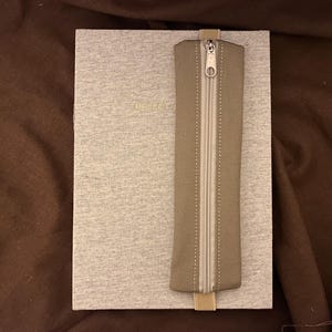Könnte beinhalten: Ein hellgraues Notizbuch mit dem Wort "BELLE" in Gold geprägt. Ein olivgrünes Federmäppchen mit weißem Reißverschluss ist mit einem beigefarbenen Gummiband am Notizbuch befestigt. Das Notizbuch und das Federmäppchen liegen auf einer braunen Oberfläche.