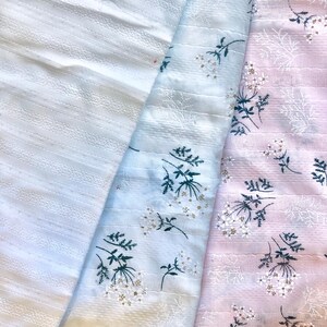 K&ouml;nnte beinhalten: Drei Stoffmuster in Wei&szlig;, Hellblau und Rosa. Der wei&szlig;e Stoff hat ein strukturiertes Streifenmuster. Die hellblauen und rosa Stoffe weisen einen Blumendruck mit wei&szlig;en Bl&uuml;ten und gr&uuml;nen Stielen auf. Die Stoffe sind gefaltet.