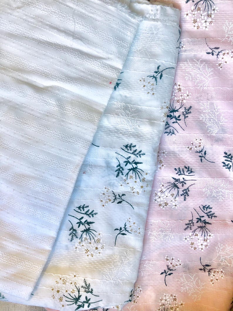 K&ouml;nnte beinhalten: Drei Stoffmuster in Wei&szlig;, Hellblau und Rosa. Der wei&szlig;e Stoff hat ein strukturiertes Streifenmuster. Die hellblauen und rosa Stoffe weisen einen Blumendruck mit wei&szlig;en Bl&uuml;ten und gr&uuml;nen Stielen auf. Die Stoffe sind gefaltet.