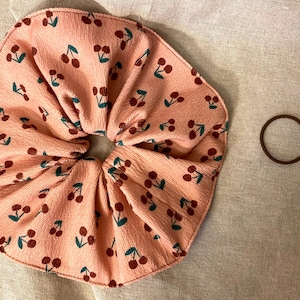 May include: A peach-colored scrunchie with a repeating pattern of dark red cherries and green leaves. A separate brown hair tie is next to the scrunchie. The scrunchie is made of a textured fabric.