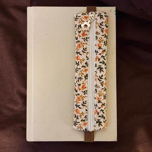 Peut inclure: Un cahier beige avec une pochette à fermeture éclair florale attachée. La pochette a une fermeture éclair blanche et une sangle marron. Le motif floral comprend des feuilles orange et vertes sur fond blanc. Le curseur de la fermeture éclair est en forme de fleur.