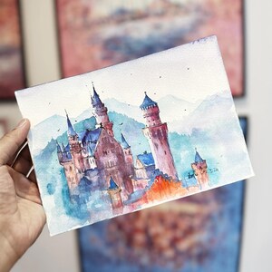 K&ouml;nnte beinhalten: Aquarellmalerei eines M&auml;rchenschlosses in Blau- und Rosat&ouml;nen. Das Schloss ist von B&auml;umen und Bergen umgeben, der Himmel ist hellblau. Das Bild ist signiert mit "Lori O'Neil 2024".
