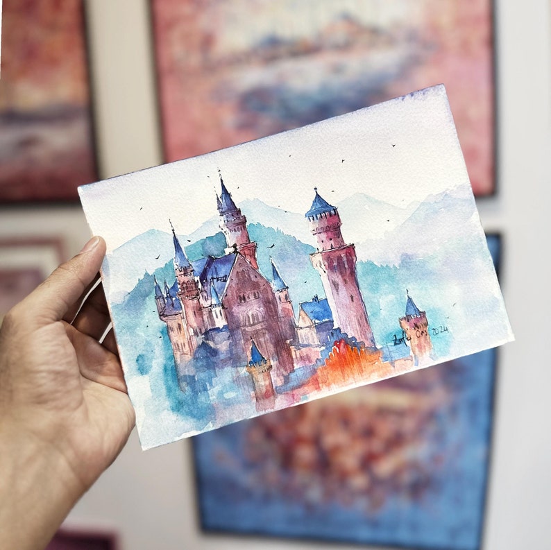K&ouml;nnte beinhalten: Aquarellmalerei eines M&auml;rchenschlosses in Blau- und Rosat&ouml;nen. Das Schloss ist von B&auml;umen und Bergen umgeben, der Himmel ist hellblau. Das Bild ist signiert mit "Lori O'Neil 2024".