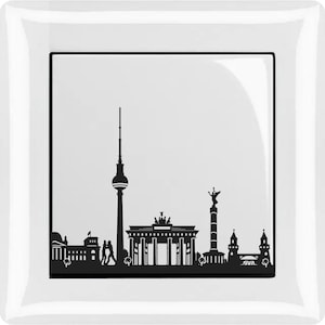 Op de afbeelding: Een witte lichtschakelaar-afdekplaat met een zwart silhouet van de skyline van Berlijn, inclusief de Brandenburger Tor, de Berliner Fernsehturm en de Siegessäule.