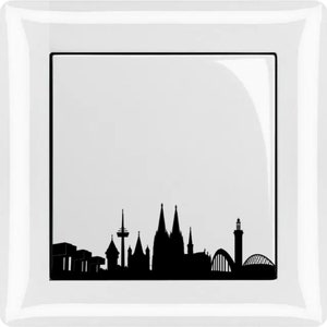 Può includere: Un piatto quadrato bianco con una silhouette nera di uno skyline cittadino. Lo skyline include edifici, un ponte e una torre.