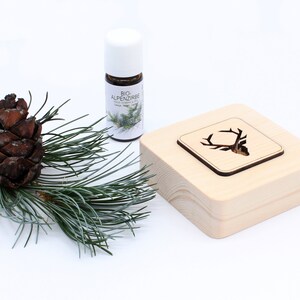 Può includere: Un diffusore in legno con un design a testa di cervo e una piccola bottiglia di olio essenziale Bio-Alpenzirbe. Il diffusore è in legno chiaro e ha una forma quadrata. La bottiglia è in vetro trasparente con un tappo bianco e contiene un liquido marrone scuro.